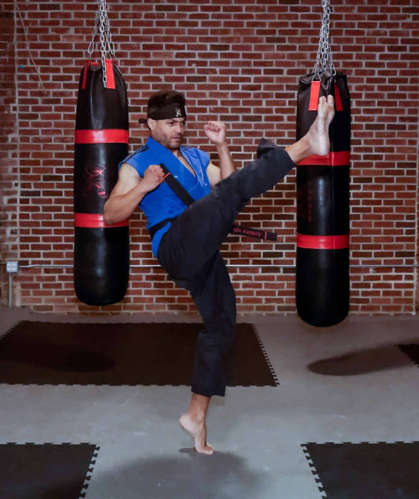 Man in blue karate gi performs high roundhouse kick toward punching bag in dimly lit brick dojo with hanging bags and mats.