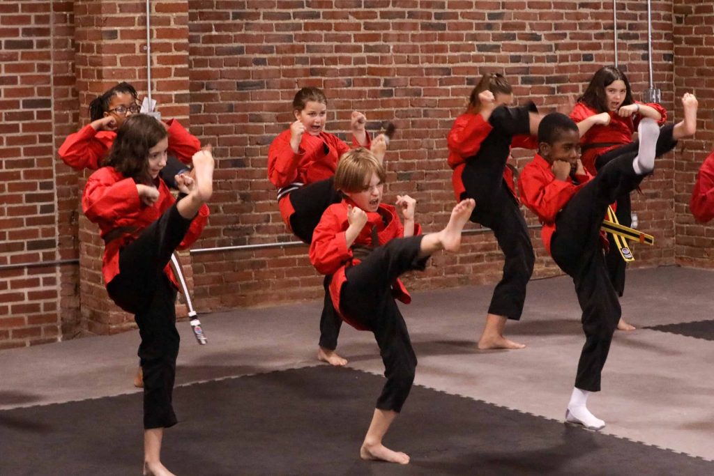 Eight kids in red karate uniforms perform synchronized high kicks in a brick dojo, fists raised and faces focused.